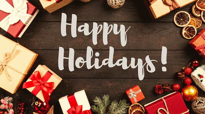 Gifts Happy Holidays Happy Holidays in a white, fun font on a dark brown table surrounded by brightly wrapped gifts and holiday ornaments