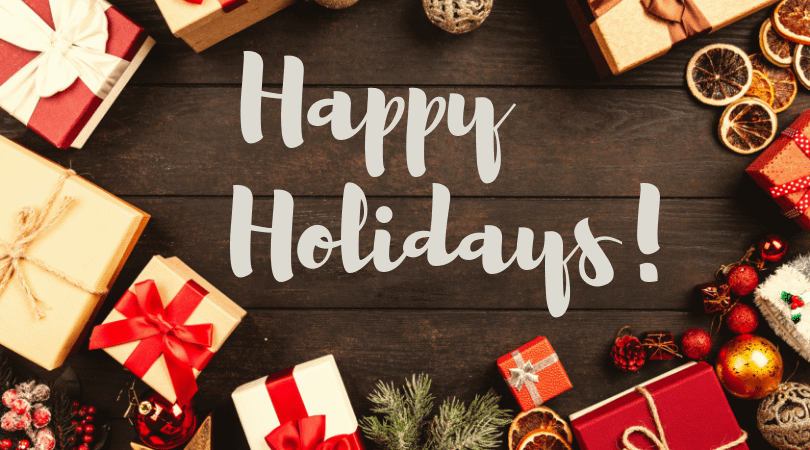 Happy Holidays in a white, fun font on a dark brown table surrounded by brightly wrapped gifts and holiday ornaments